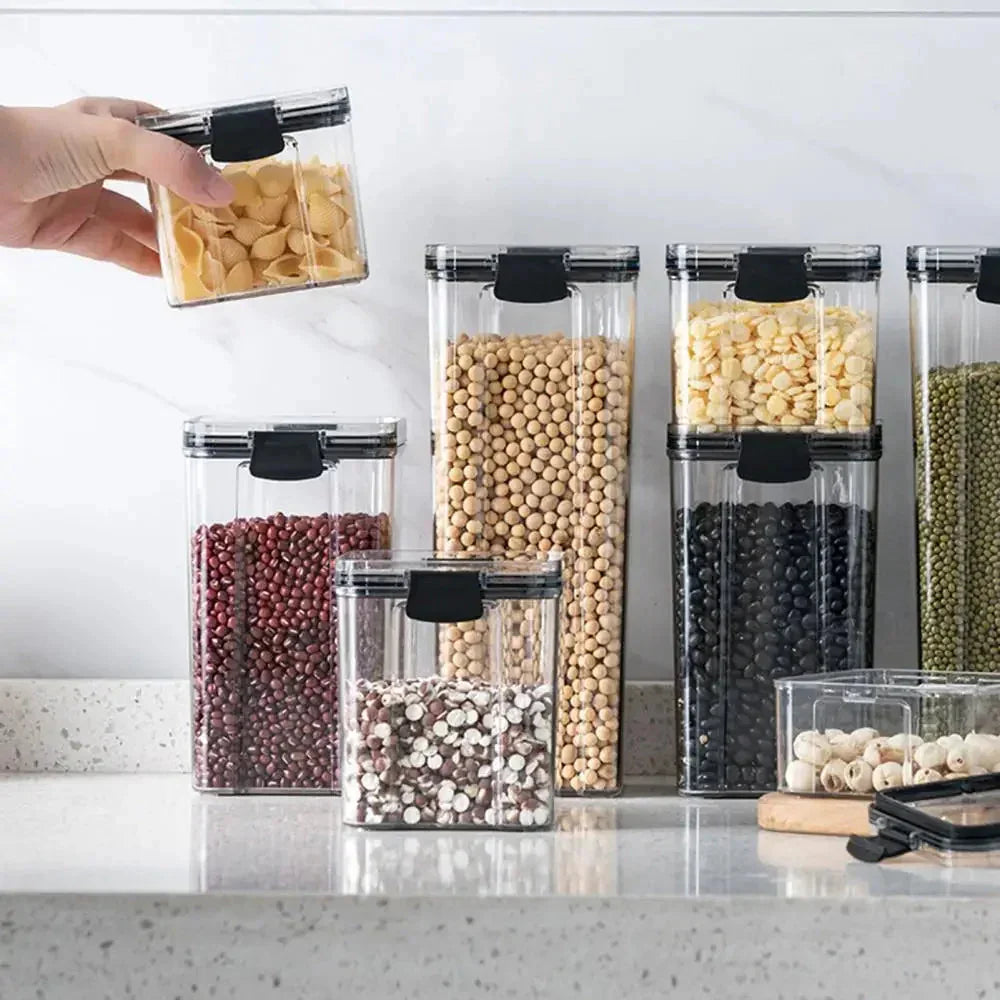 Clear kitchen storage containers with black lids holding pasta, beans, and dried goods on a countertop.